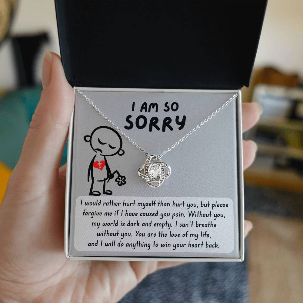 A hand holds an open jewelry box containing the Sorry-Dark And Empty - Love Knot Necklace, a heart-shaped pendant adorned with cubic zirconia crystals on a chain. The box has a message of apology and love printed inside, highlighting this necklace as the perfect gift available in its white gold or yellow gold finish.