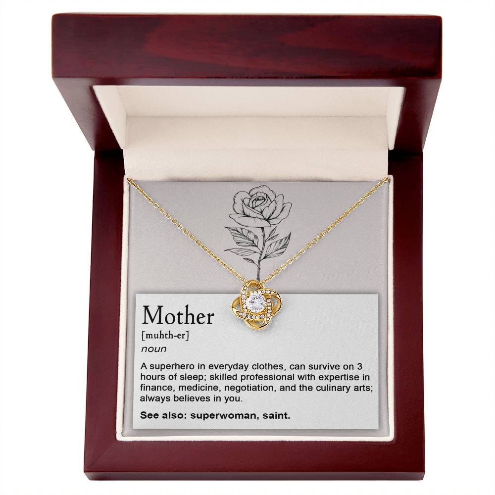 A "To Mom, Superhero - Love Knot Necklace" with a white gold finish and a pendant featuring a central gem adorned with cubic zirconia crystals is displayed in an open wooden box. Inside the lid, an engraved rose and a definition of "Mother," including attributes and synonyms, are visible.