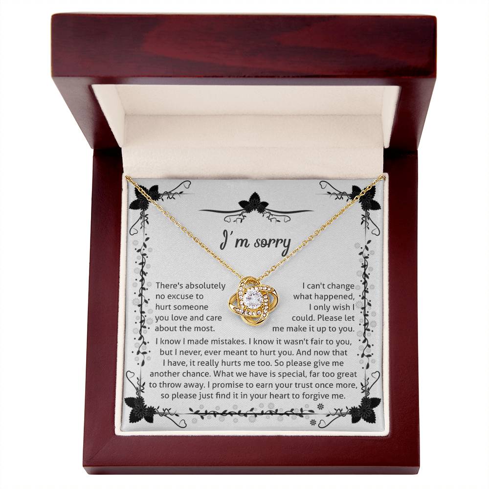 The Sorry-No Excuse - Love Knot Necklace, with a white gold finish, showcases a delicate knot design on a chain adorned with cubic zirconia crystals. The pendant is presented on a box featuring an "I'm sorry" message that expresses regret and seeks forgiveness, framed by a charming floral border at the top.