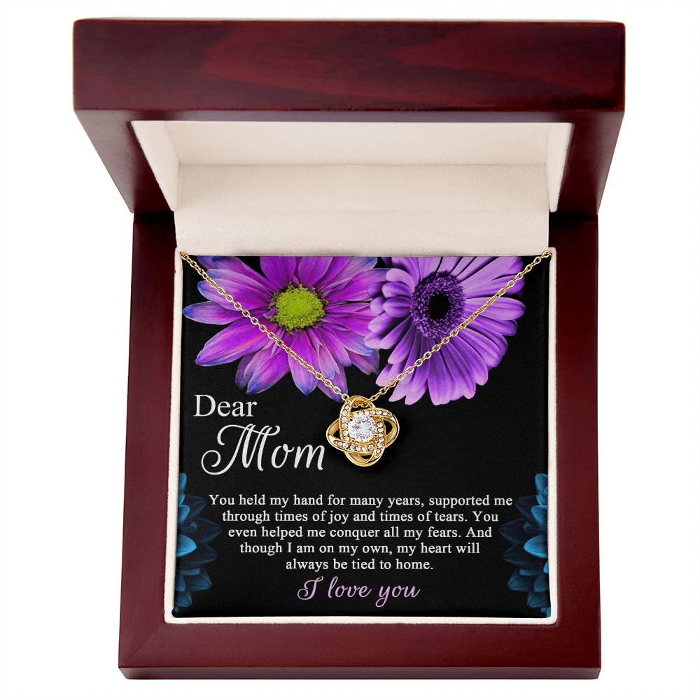 A "To Mom, Held My Hand - Love Knot Necklace" adorned with cubic zirconia crystals, displayed in an open wooden box with purple flowers and an inscription dedicated to "Mom.