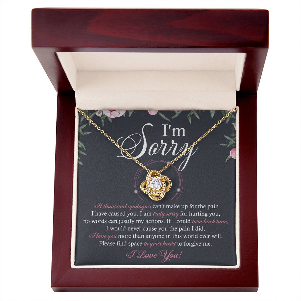 A box containing the "Sorry-World Ever Will - Love Knot Necklace," featuring a central pendant adorned with cubic zirconia crystals. The box lid displays text apologizing and seeking forgiveness, framed by a decorative floral border.