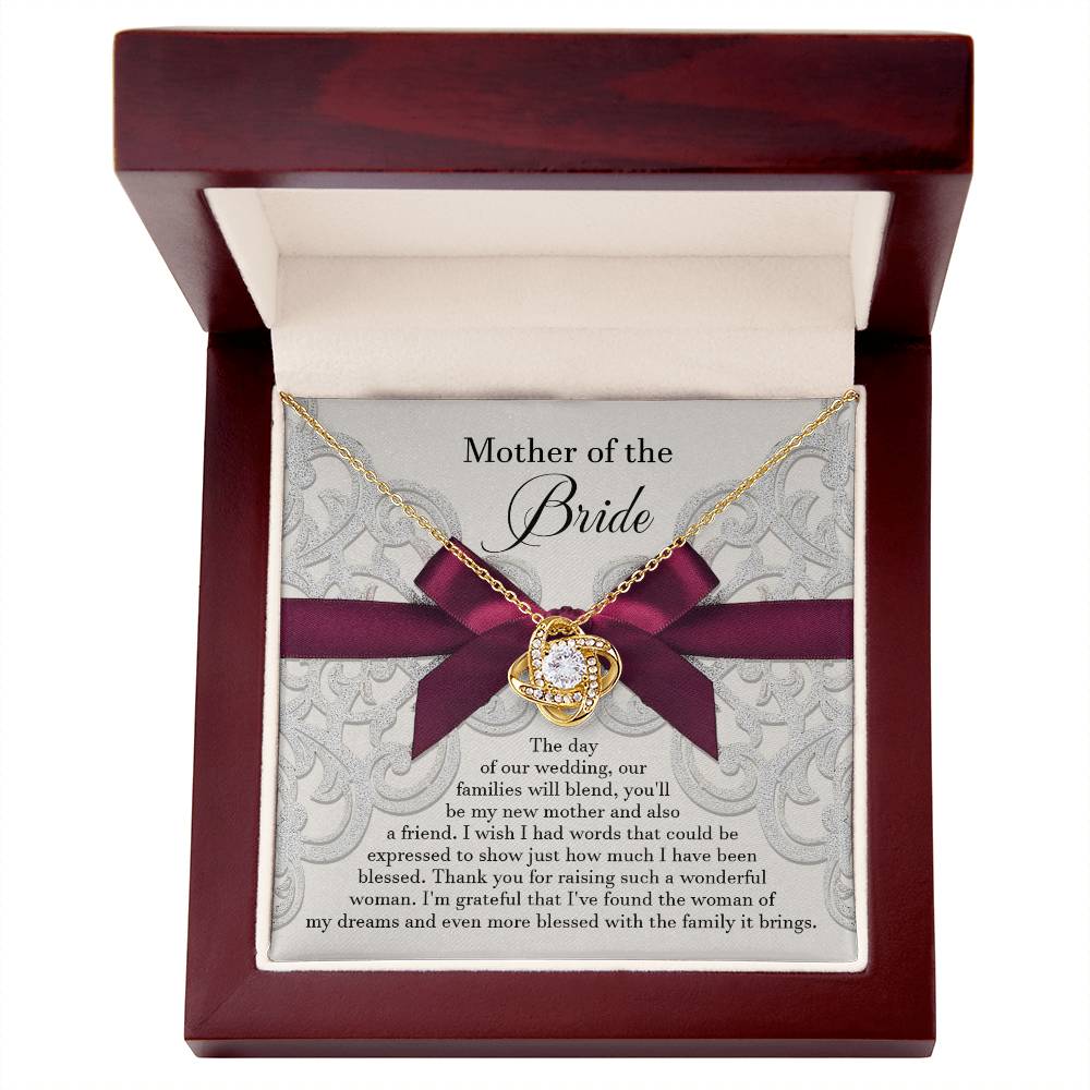 A To Bride's Mom, My New Mother - Love Knot Necklace with a heart pendant, crafted from 14k white gold and adorned with cubic zirconia crystals, is displayed on a decorative card with a maroon ribbon. The card reads, "Mother of the Bride" and contains a heartfelt message of gratitude.