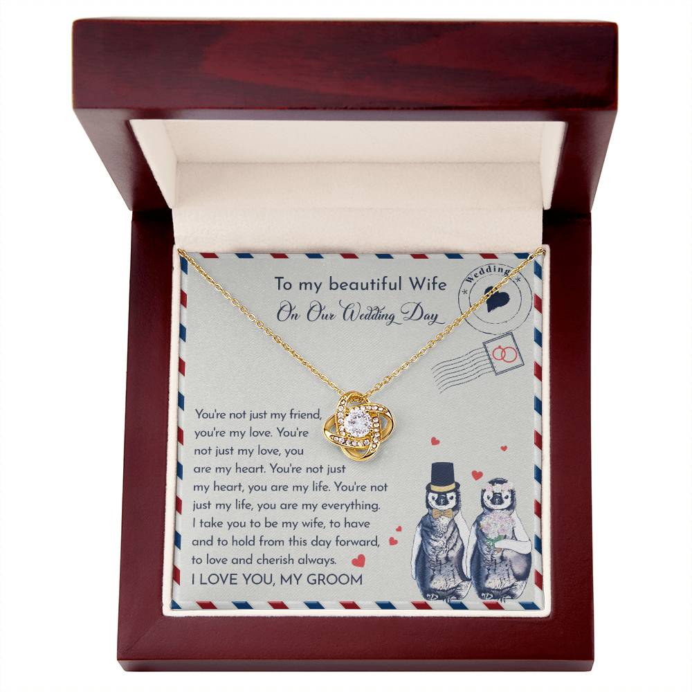 Close-up of a To Wife Wedding, Love And Cherish - Love Knot Necklace with cubic zirconia crystals on a card. The 14k white gold pendant is heart-shaped, and the card has a message for a wife on the wedding day. Two penguins, dressed as a bride and groom, are illustrated at the bottom.