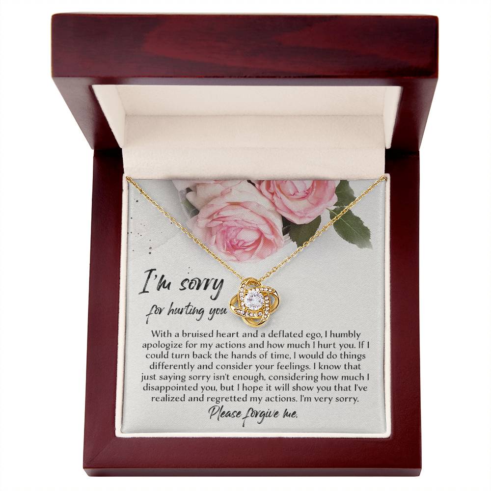 The "Sorry-Regretted My Actions - Love Knot Necklace," crafted in 14k white gold with an intricate pendant adorned with cubic zirconia crystals, is displayed against a backdrop featuring a printed apology note surrounded by roses. The note expresses regret and asks for forgiveness for past actions.