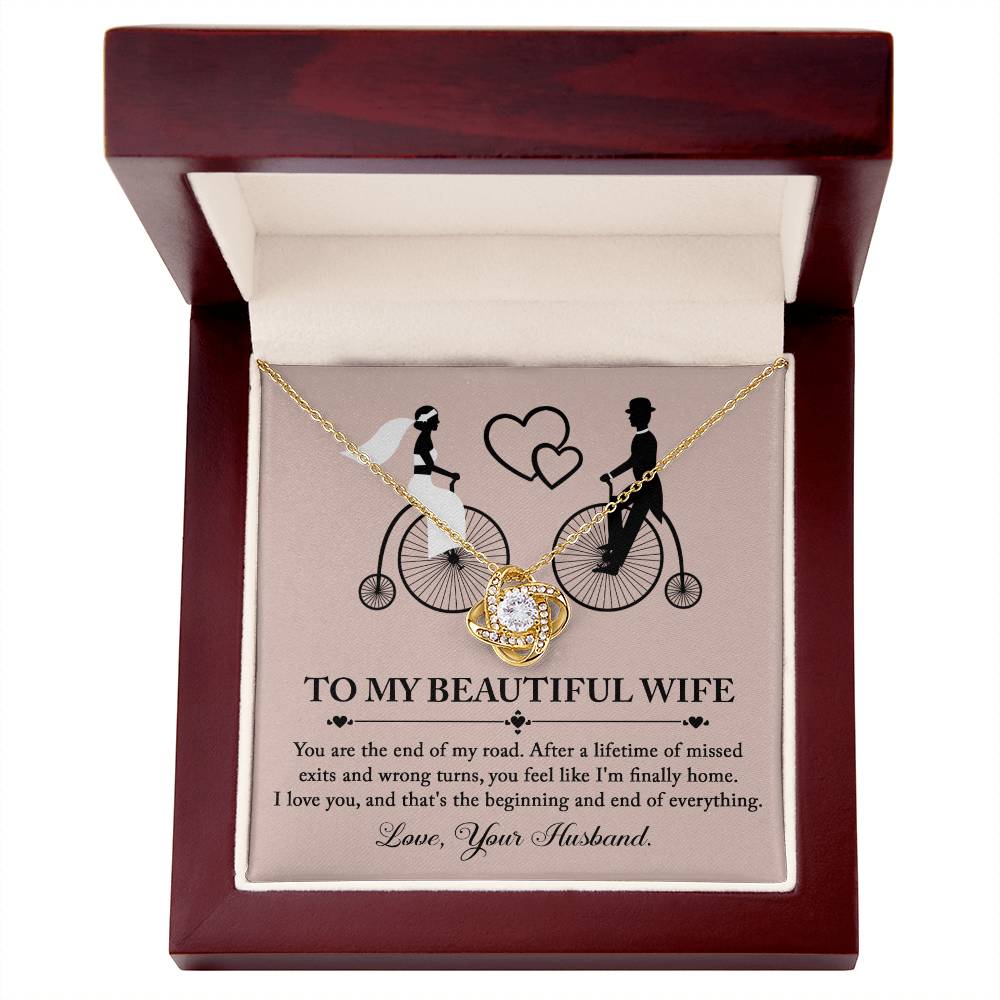 To Wife, I'm Finally Home - Love Knot Necklace with intertwined circle pendant on a card featuring an illustration of a couple on bicycles. The necklace, adorned with cubic zirconia crystals and a stunning gold finish, comes with a card that reads: 'To my beautiful wife' followed by a romantic message and ending with 'Love, Your Husband.'