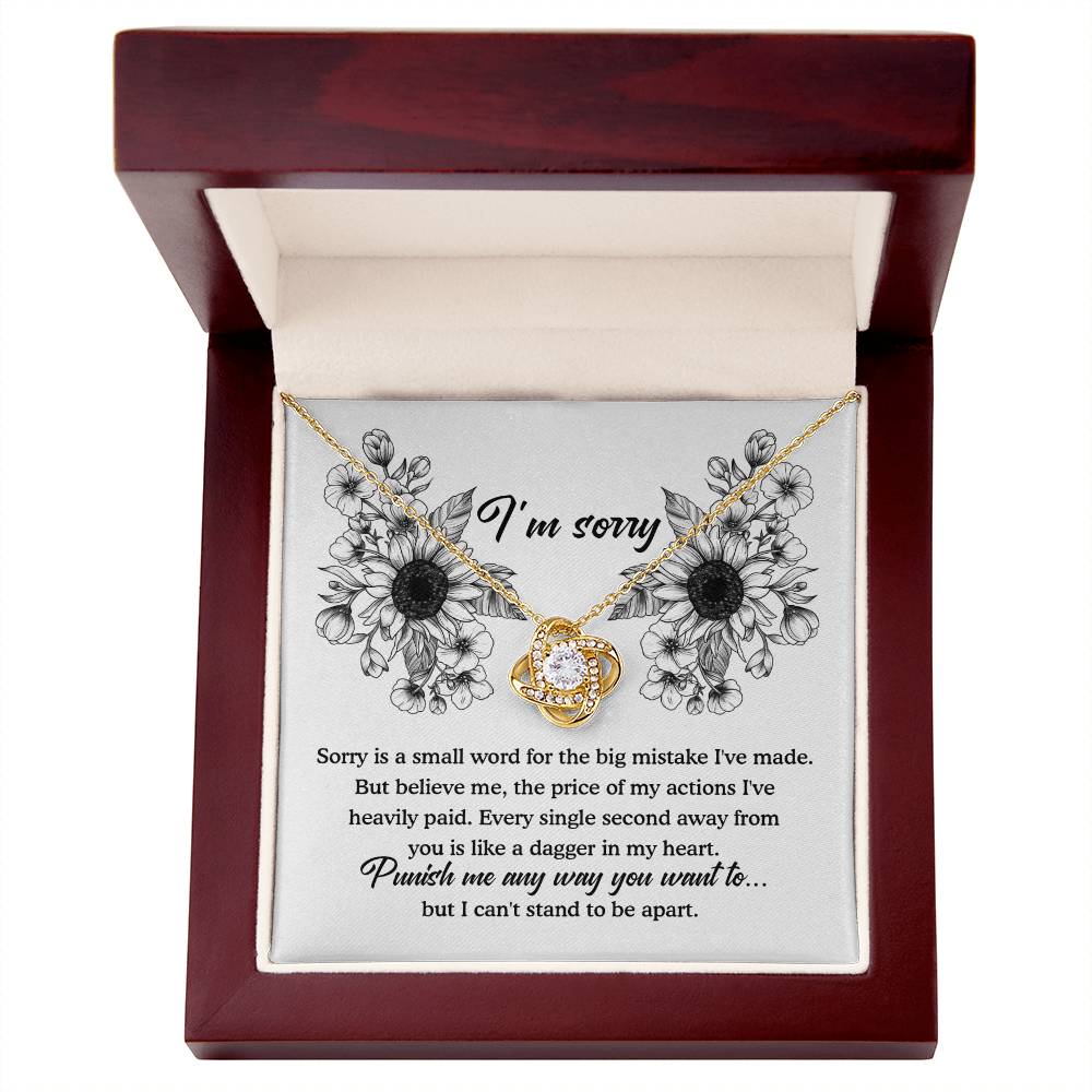 The Sorry-Small Word - Love Knot Necklace with a gold finish is elegantly showcased in an open wooden box. Inside the lid, there's a heartfelt apology note adorned with a floral design around the words "I'm sorry," accompanied by a personalized message expressing regret and longing for the recipient's forgiveness.