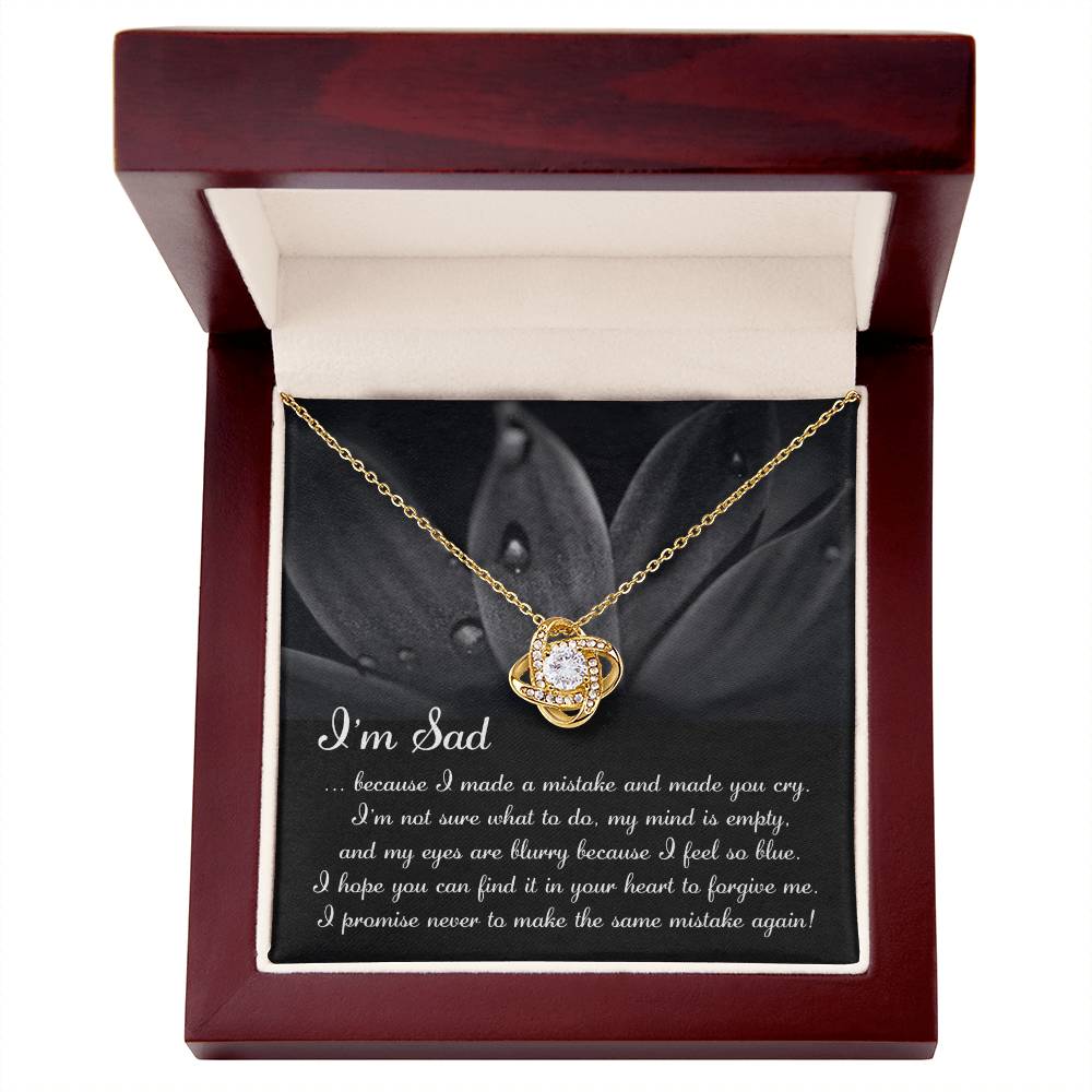 The "Sorry-Feel So Blue - Love Knot Necklace," featuring a stunning knot pendant adorned with cubic zirconia crystals, is displayed over a card with an apologetic message starting with "I'm Sad" and set against a floral background. This personalized gift beautifully combines elegance and heartfelt sentiment.