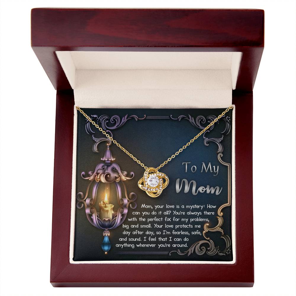 The "To Mom, Love Is Mystery - Love Knot Necklace" is beautifully showcased in an open wooden jewelry box. Inside the lid, there's a card adorned with a decorative lamp design and a message that reads, "To My Mom," along with a heartfelt note. This gold necklace features cubic zirconia, adding an extra touch of elegance.