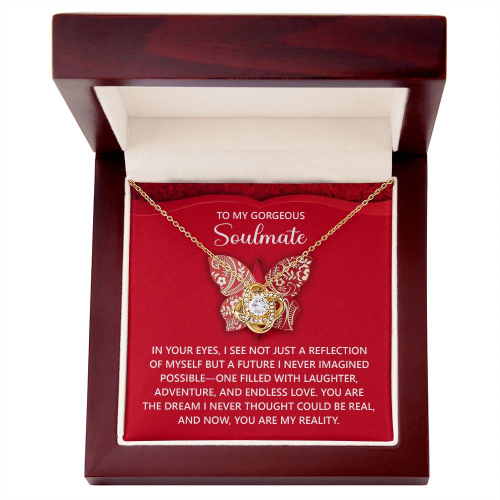 The Soulmate-My Reality Love Knot Necklace, featuring cubic zirconia crystals, is nestled in an open red and white jewelry box with a romantic message. Its yellow gold finish enhances the elegance of this stunning piece.