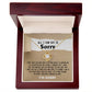 A **Sorry-Keep Me Apart - Love Knot Necklace** with a circular pendant adorned with cubic zirconia crystals is displayed in an open wooden box. Inside the box lid, a printed apology message is visible that emphasizes hope for forgiveness and expresses regret.