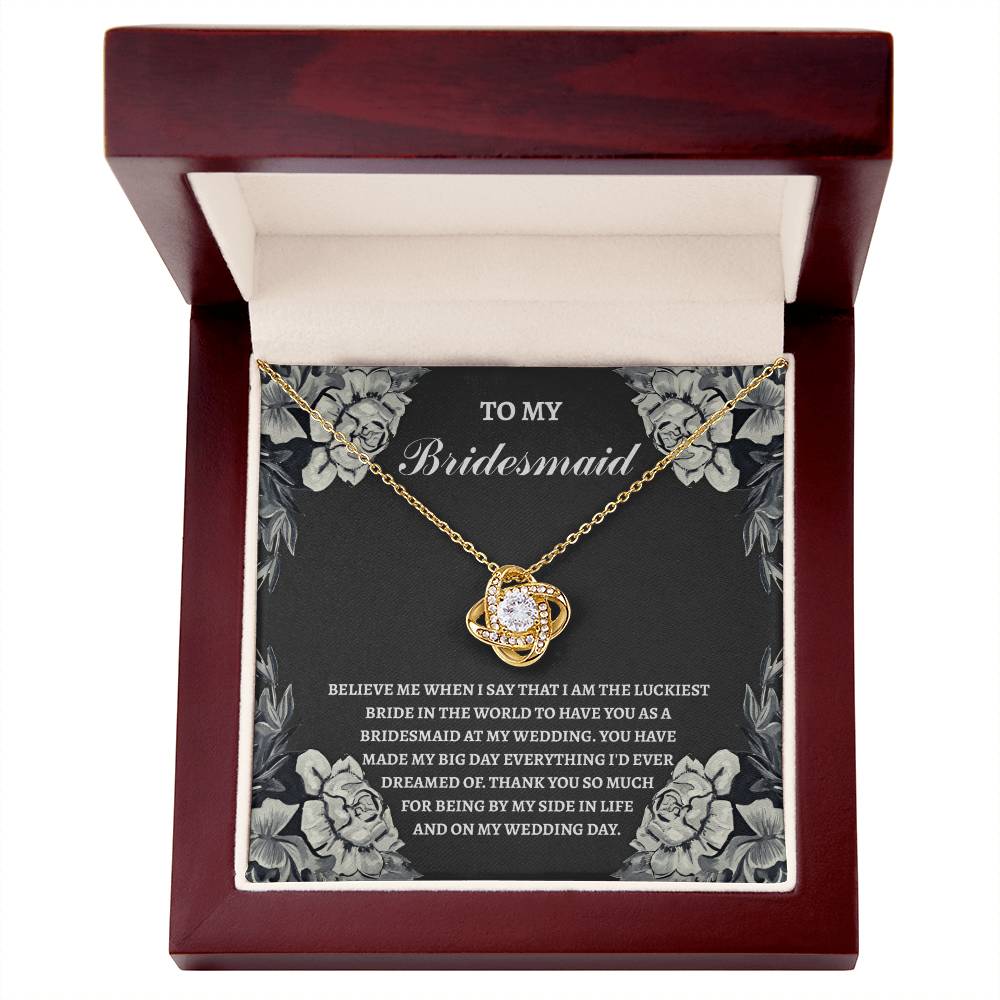 A Love Knot Necklace in gold with an ornate pendant is elegantly presented in a wooden box. Inside, the text reads: "To My Bridesmaid" and includes a heartfelt message expressing gratitude for her support at the wedding. The product name is "To Bridesmaid, The Luckiest Bride - Love Knot Necklace.