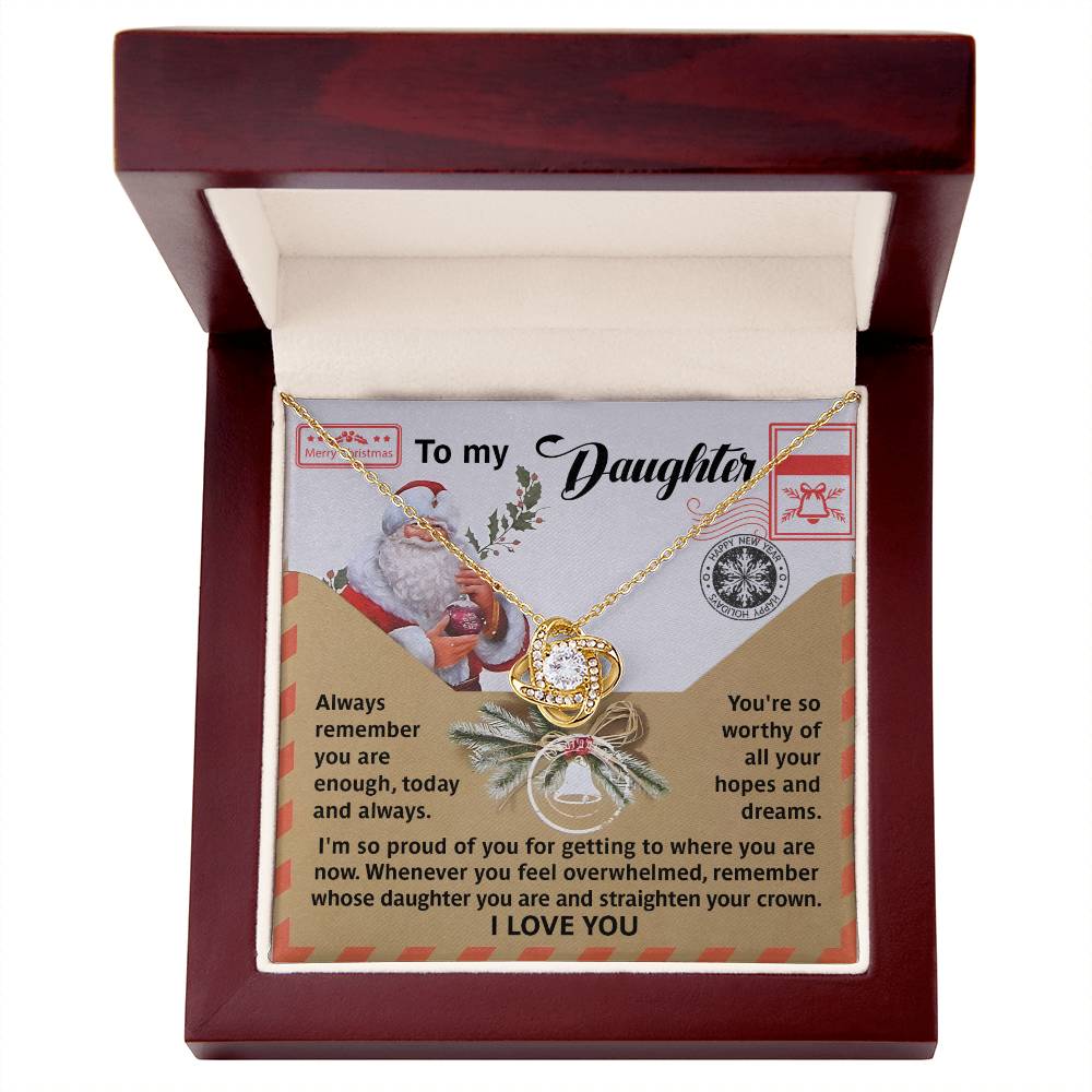 Inside the open wooden jewelry box lies a stunning Daughter-All Your Hopes - Love Knot Necklace adorned with cubic zirconia crystals. The necklace rests on a card featuring a heartfelt message to a daughter, surrounded by festive holiday graphics like Santa Claus and twinkling decorations, all in an elegant gold finish.