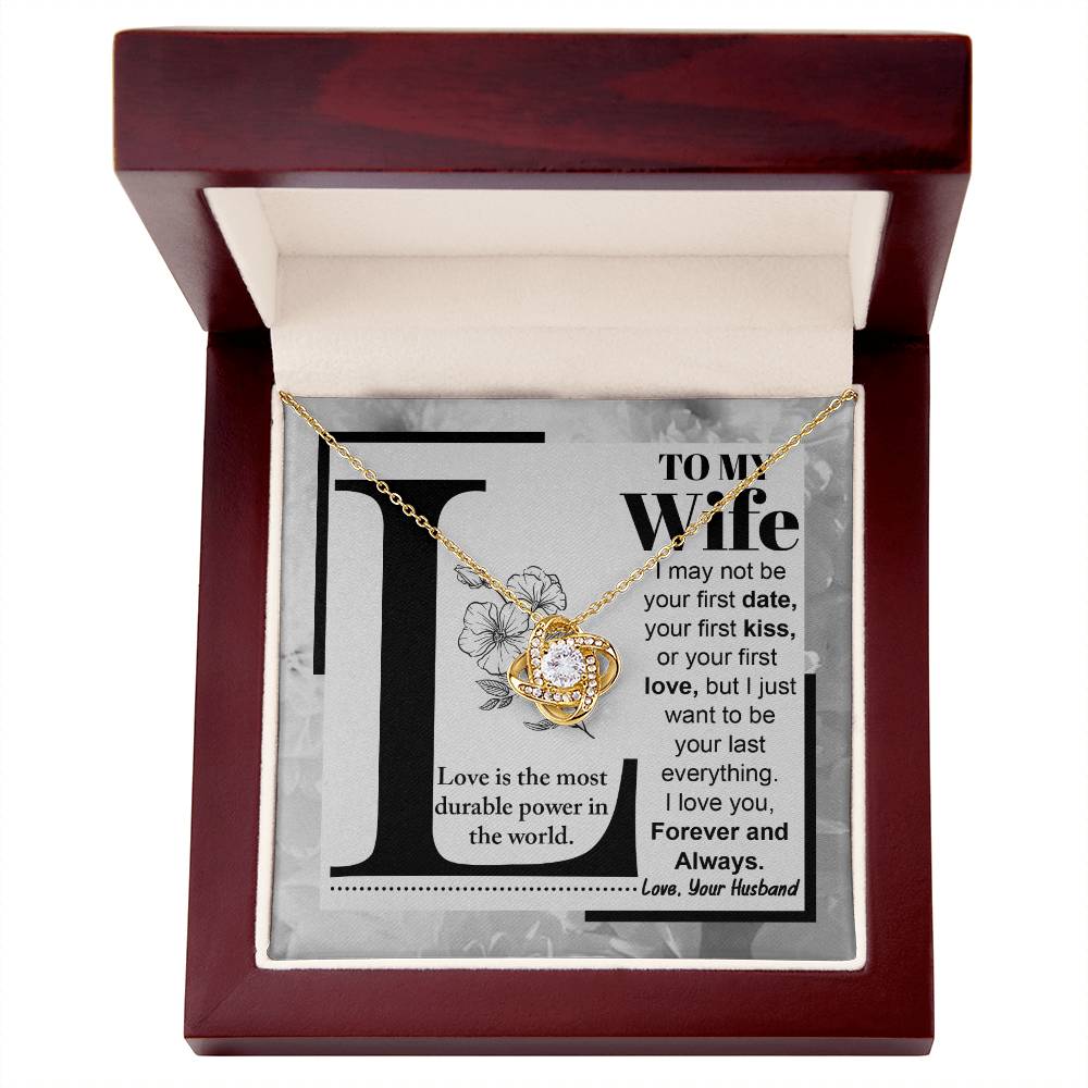 A "to Wife, Love Is - Love Knot Necklace" with a knot design is displayed on a card featuring a printed message to a wife about lasting love, signed from a husband. The piece sparkles with cubic zirconia crystals and boasts an elegant gold finish.