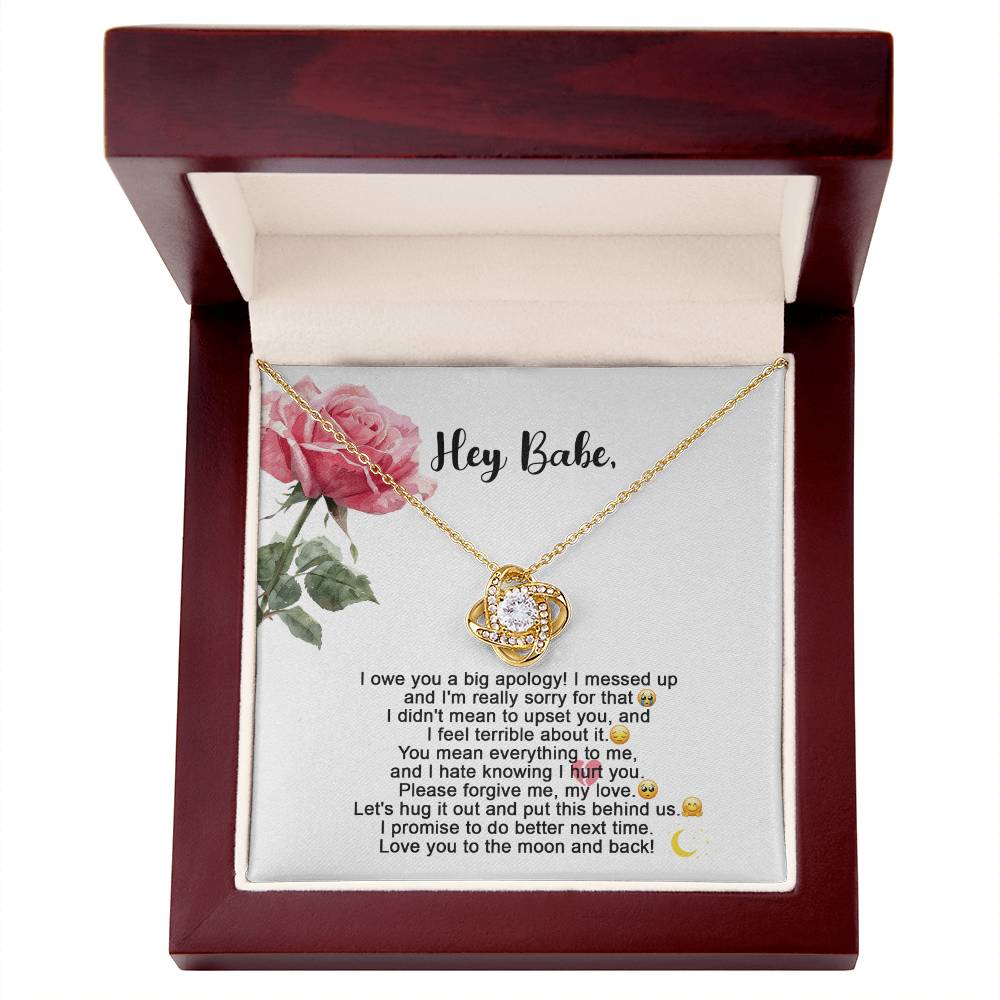 The "Sorry-Hug It Out - Love Knot Necklace" features an 18k yellow gold pendant adorned with small cubic zirconia crystals. It comes on a card decorated with roses, accompanied by the message "Hey Babe" and an apology note.