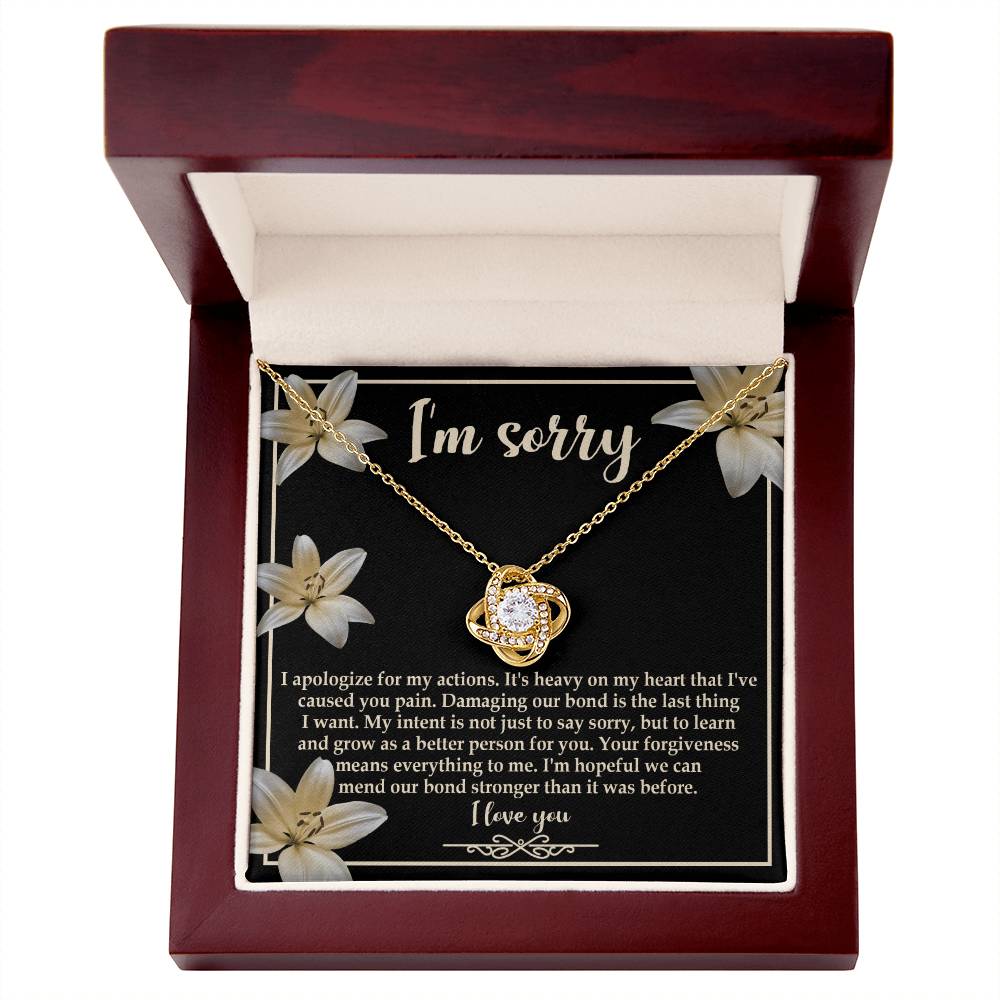 The "Sorry-The Last Thing - Love Knot Necklace" showcases a love knot pendant against a striking black background adorned with white flowers. It's accompanied by an apology message that reads, "I'm sorry. I apologize for my actions... I'm hopeful we can mend our bond stronger," and is beautifully finished with white gold and shimmering cubic zirconia crystals.