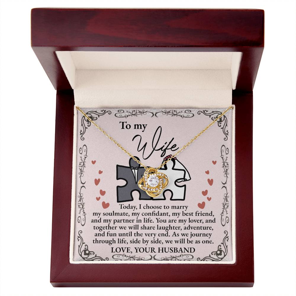 A "To Wife, Be As One - Love Knot Necklace" with intertwined hearts is placed on a card adorned with cubic zirconia crystals, expressing love and commitment from a husband to his wife. The card features a heart and puzzle piece design, available in white gold or yellow gold finish.