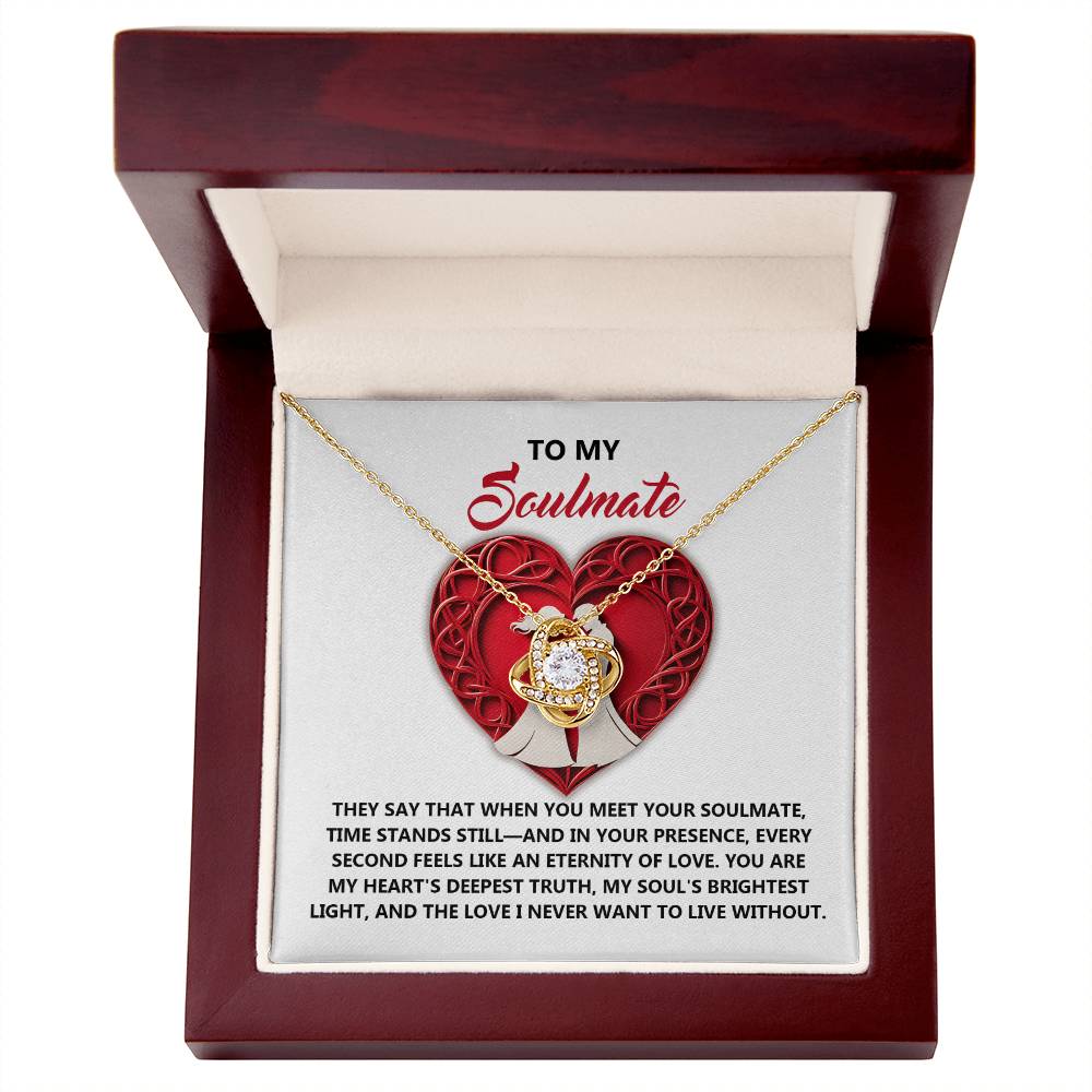 The Soulmate-Deepest Truth Love Knot Necklace comes in a box with a romantic message against a red and white backdrop. Its heart-shaped pendant with a shimmering gold finish adds elegance, making it an ideal gift for your special someone.