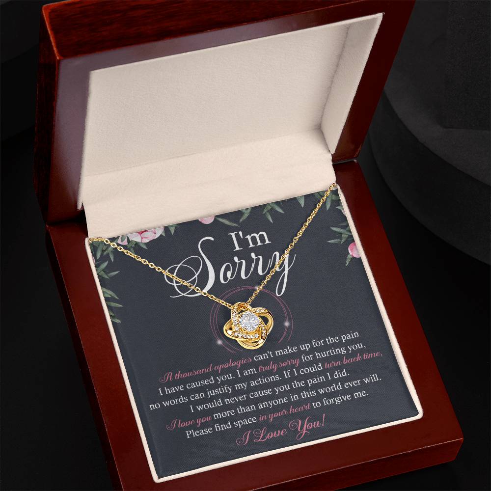 A box containing the "Sorry-World Ever Will - Love Knot Necklace," featuring a central pendant adorned with cubic zirconia crystals. The box lid displays text apologizing and seeking forgiveness, framed by a decorative floral border.