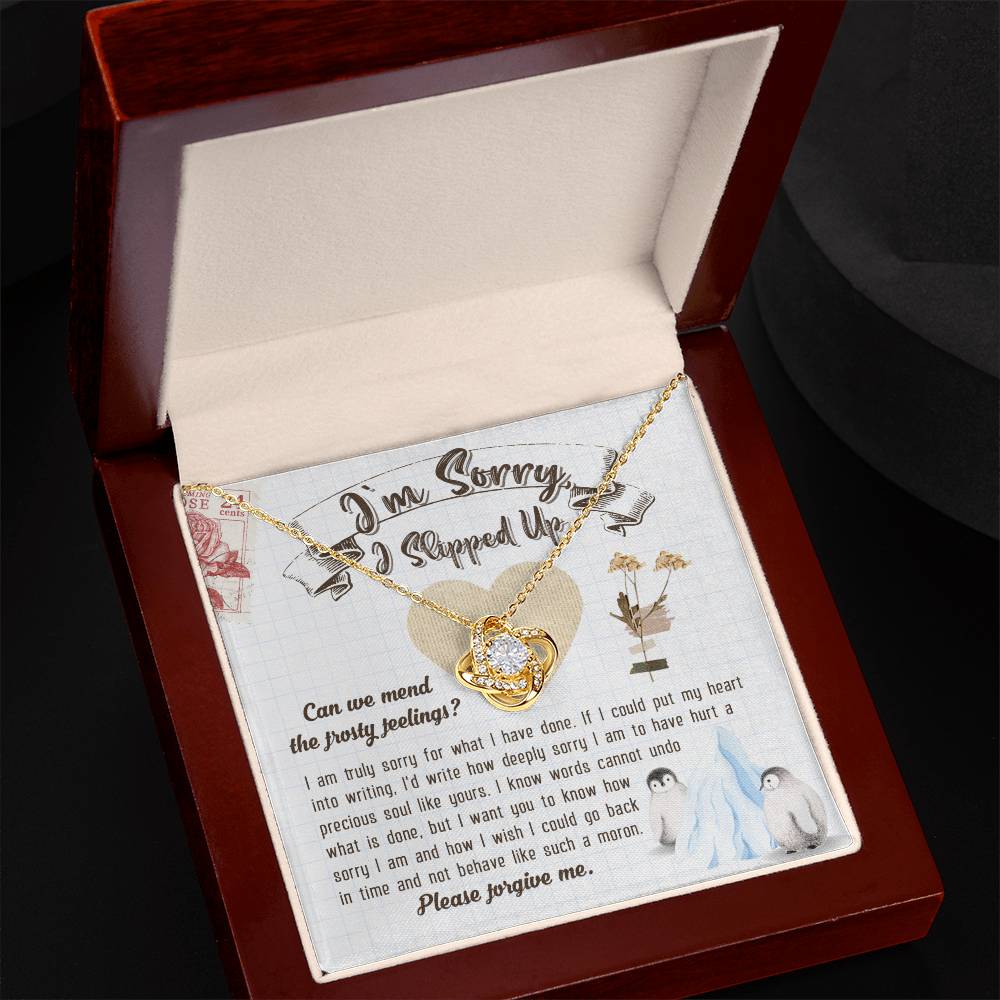 The Sorry-Precious Soul - Love Knot Necklace, featuring an intertwined heart pendant adorned with cubic zirconia crystals, is beautifully displayed in a box. The box comes with an apology note and charming illustrations of flowers, mail stamps, and penguins on a grid-patterned background.