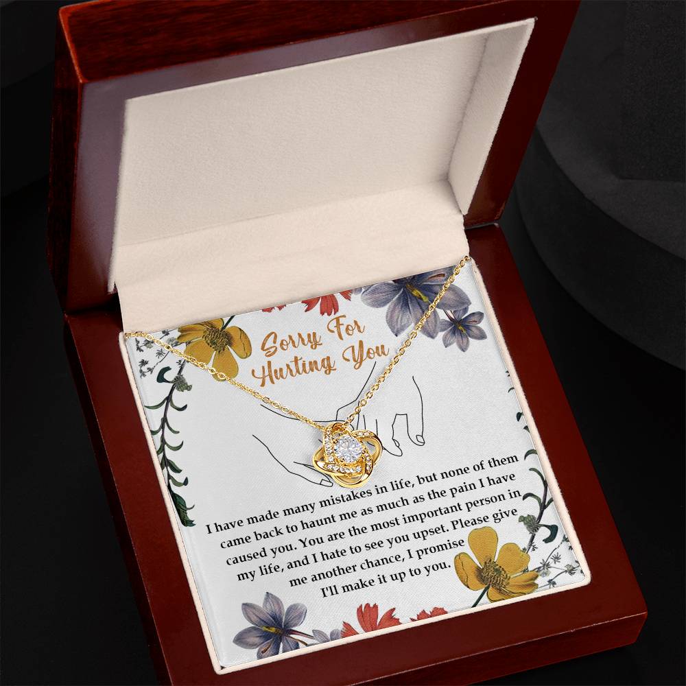 The Sorry-Haunt Me - Love Knot Necklace is a silver piece featuring an intertwined heart and infinity symbol adorned with cubic zirconia crystals, elegantly presented against a floral card that reads: "Sorry For Hurting You" with an apology message below.