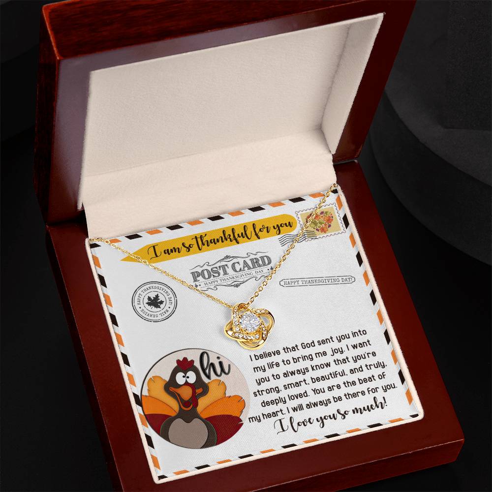 An open jewelry box reveals the Thanksgiving-The Beat - Love Knot Necklace, featuring a gold finish and charm with intertwined hearts. Inside, a postcard-like card displays a Thanksgiving message, cartoon turkey, and floral design.