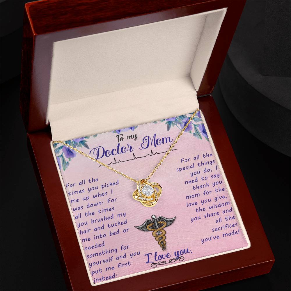 A "To My Doctor Mom, For All The Times You Picked Me Up" Love Knot Necklace adorned with cubic zirconia crystals and a heart and cross pendant is displayed inside a red jewelry box, including a message for a mother who is also a doctor, expressing gratitude.