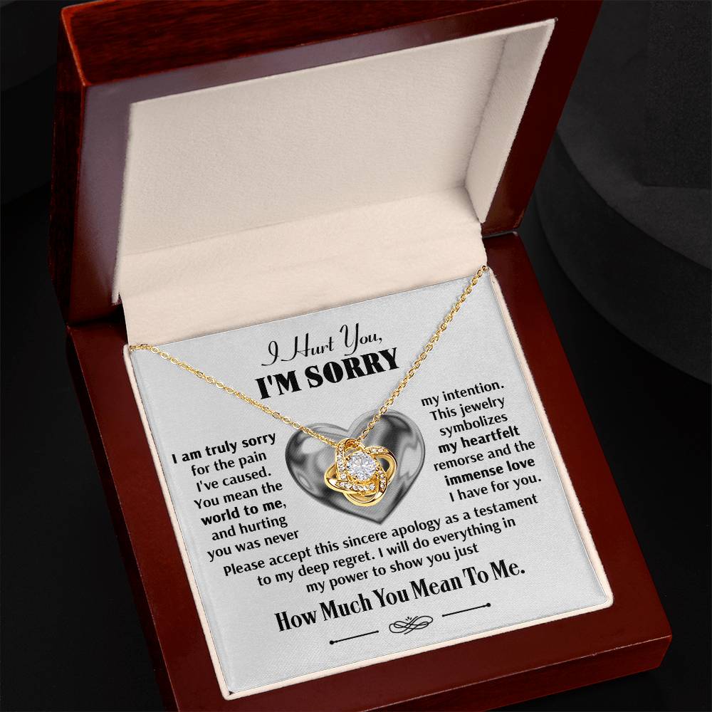 The Sorry-Sincere Apology - Love Knot Necklace, a gold heart-shaped pendant adorned with cubic zirconia crystals, is elegantly displayed in an open wooden box. It comes with a card that reads "I Hurt You, I'M SORRY" and includes an apology message underneath.