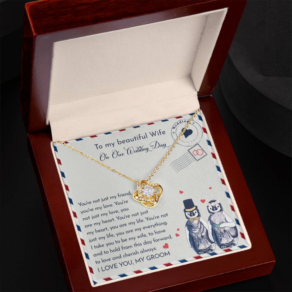 Close-up of a To Wife Wedding, Love And Cherish - Love Knot Necklace with cubic zirconia crystals on a card. The 14k white gold pendant is heart-shaped, and the card has a message for a wife on the wedding day. Two penguins, dressed as a bride and groom, are illustrated at the bottom.