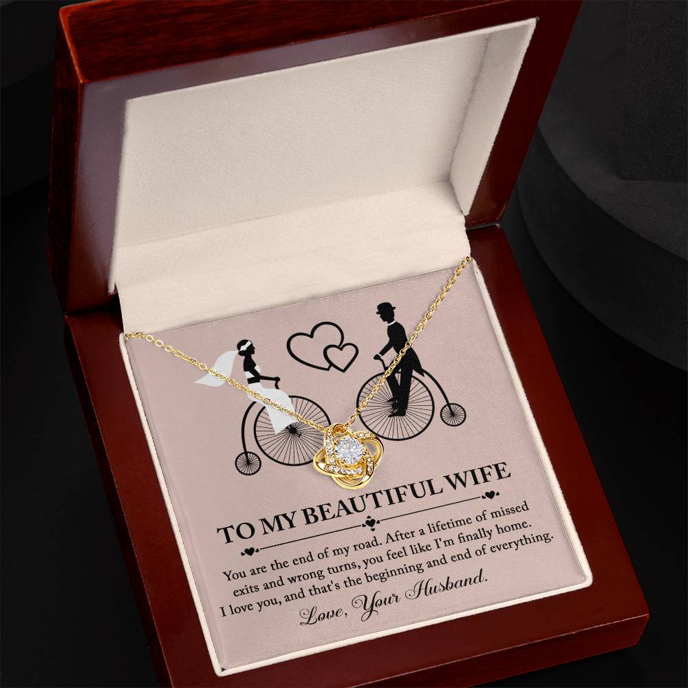 To Wife, I'm Finally Home - Love Knot Necklace with intertwined circle pendant on a card featuring an illustration of a couple on bicycles. The necklace, adorned with cubic zirconia crystals and a stunning gold finish, comes with a card that reads: 'To my beautiful wife' followed by a romantic message and ending with 'Love, Your Husband.'