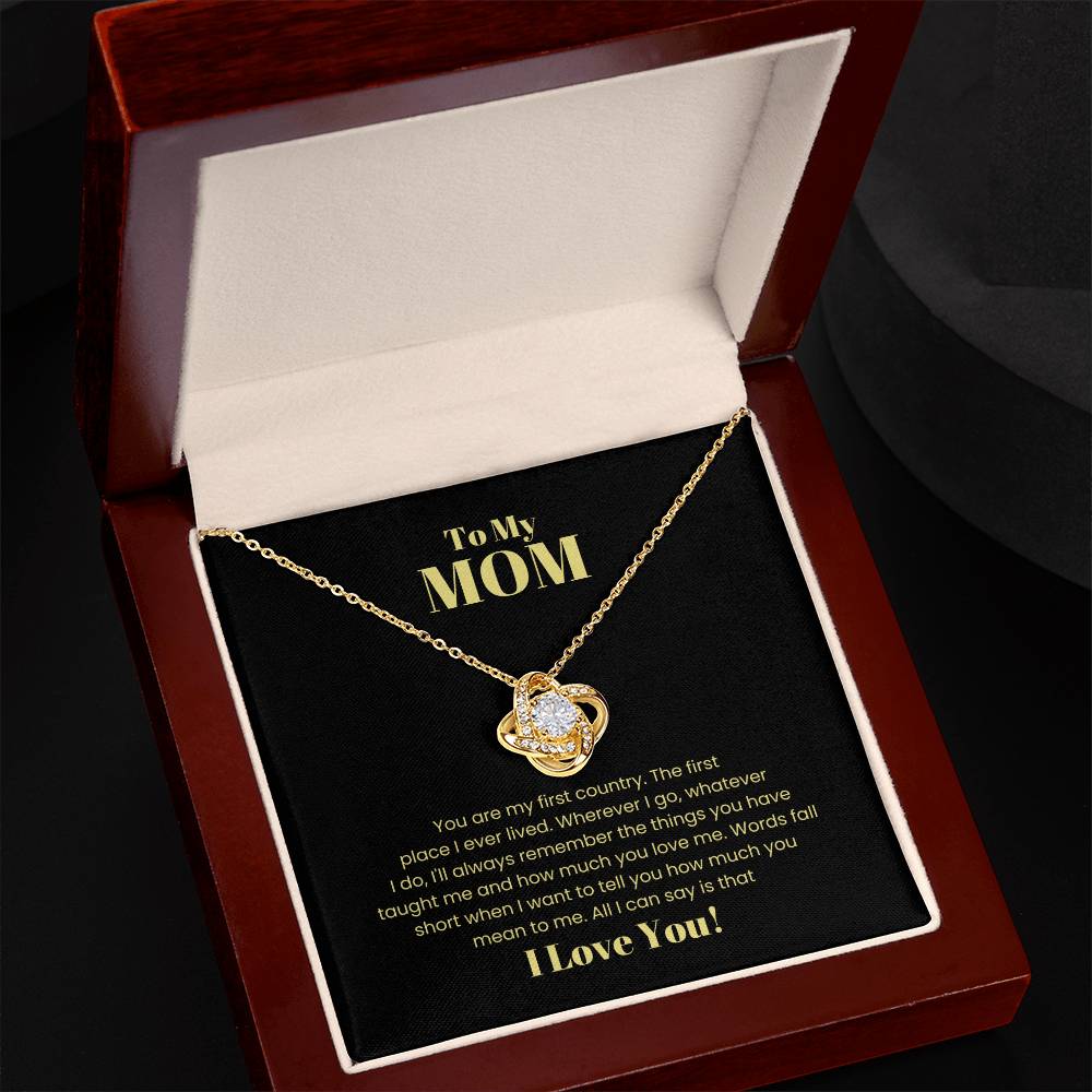 The "To My Mom, You Are My First Country - Love Knot Necklace" is displayed in an open gift box. The heart-shaped pendant, adorned with sparkling cubic zirconia crystals, is expertly crafted from 14k white gold. The inside lid features an inscription dedicated to "MOM," expressing love and gratitude.