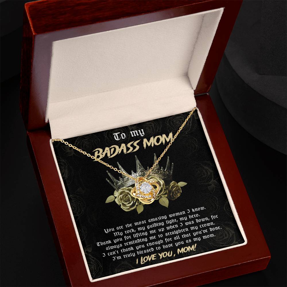 A gift box showcasing the To Mom, Straighten My Crown - Love Knot Necklace adorned with cubic zirconia crystals. The interior cover includes a message reading "To my badass mom" and a heartfelt note written below, making it the perfect personalized gift.