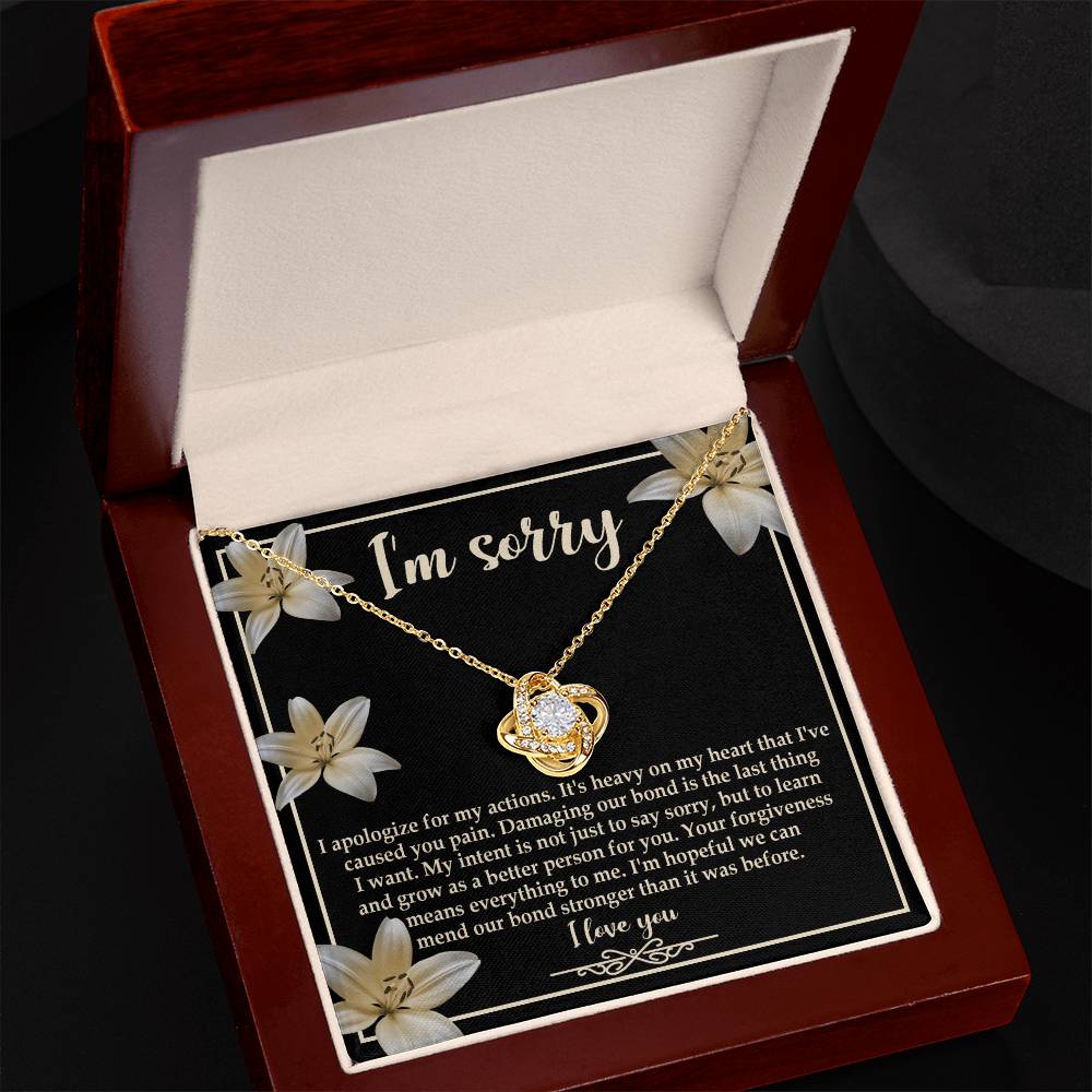 The "Sorry-The Last Thing - Love Knot Necklace" showcases a love knot pendant against a striking black background adorned with white flowers. It's accompanied by an apology message that reads, "I'm sorry. I apologize for my actions... I'm hopeful we can mend our bond stronger," and is beautifully finished with white gold and shimmering cubic zirconia crystals.