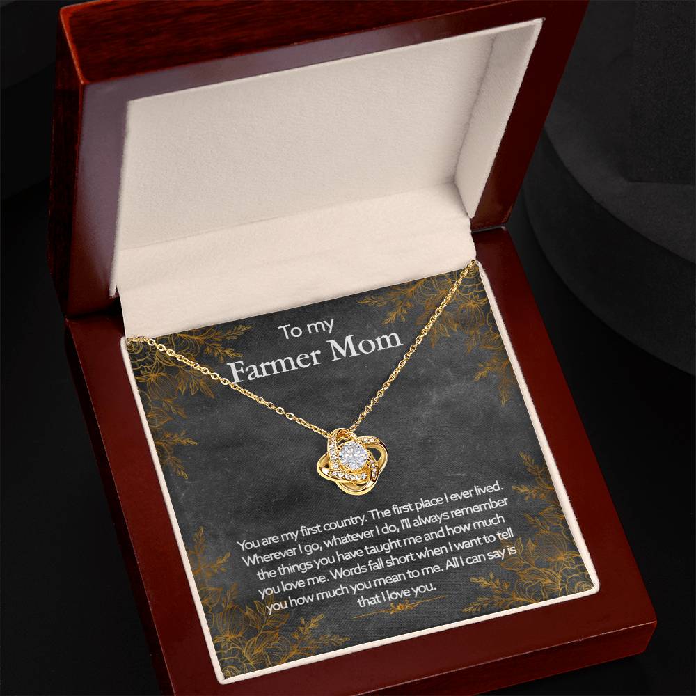 An open jewelry box containing a gold To My Farmer Mom, You Are My First Country - Love Knot Necklace with a cubic zirconia crystals pendant, presented on a card with a heartfelt message dedicated to a "farmer mom.