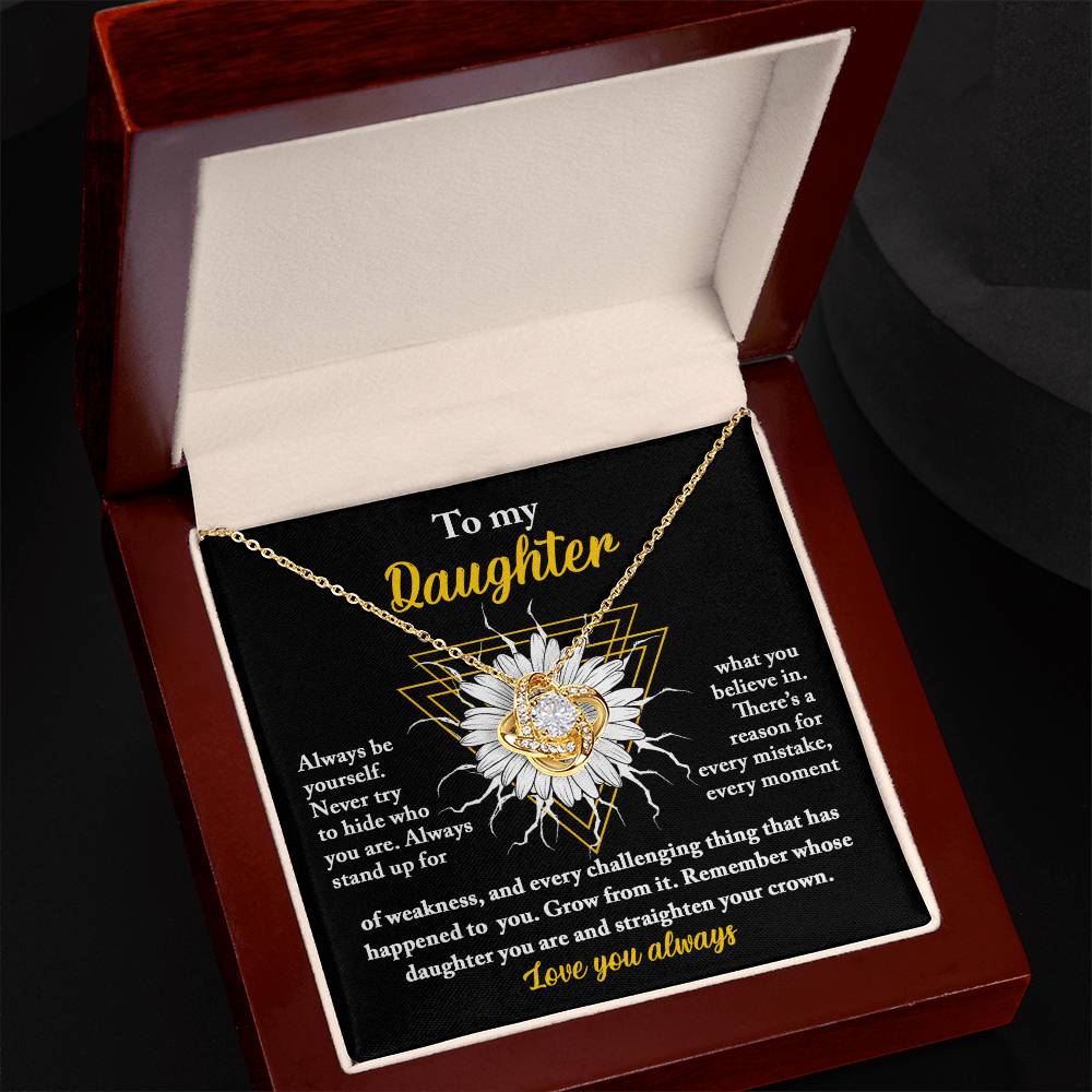 The "To Daughter, Be Yourself - Love Knot Necklace" features a heart-shaped pendant set with cubic zirconia crystals, elegantly displayed on a yellow gold chain. This stunning piece comes in an open red velvet-lined box. The inscription reads: "To my Daughter," followed by an inspiring message about strength and courage. The necklace is showcased against an intricately designed background.