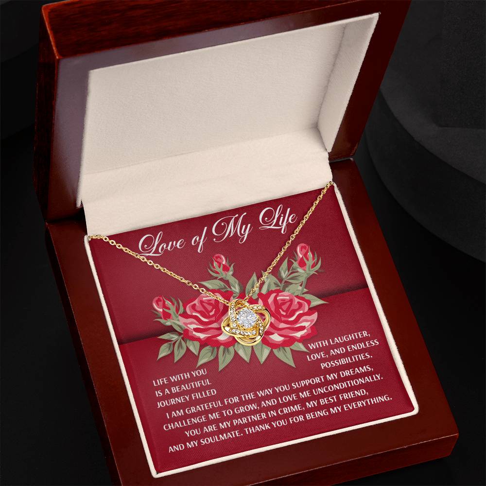 The Soulmate-Endless Possibilities Love Knot Necklace, nestled in a box, features a heart-shaped pendant with cubic zirconia crystals against the "Love of My Life" text and red roses. It is available in white or yellow gold finish.