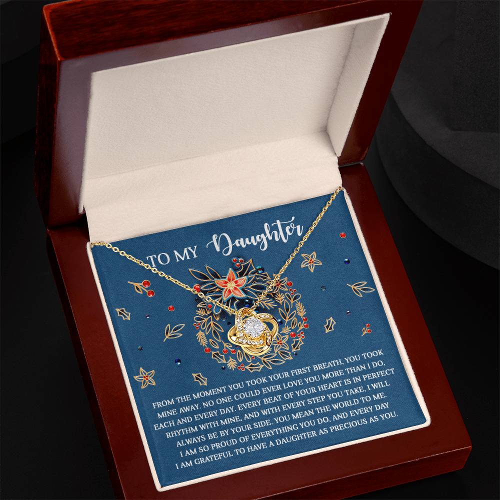 A Daughter-Perfect Rhythm - Love Knot Necklace with a heart pendant rests in an open box. Inside the lid, a sentimental message reads "To My Daughter." The cushioned box, featuring a blue floral design, cradles this personalized gift adorned with sparkling cubic zirconia crystals.