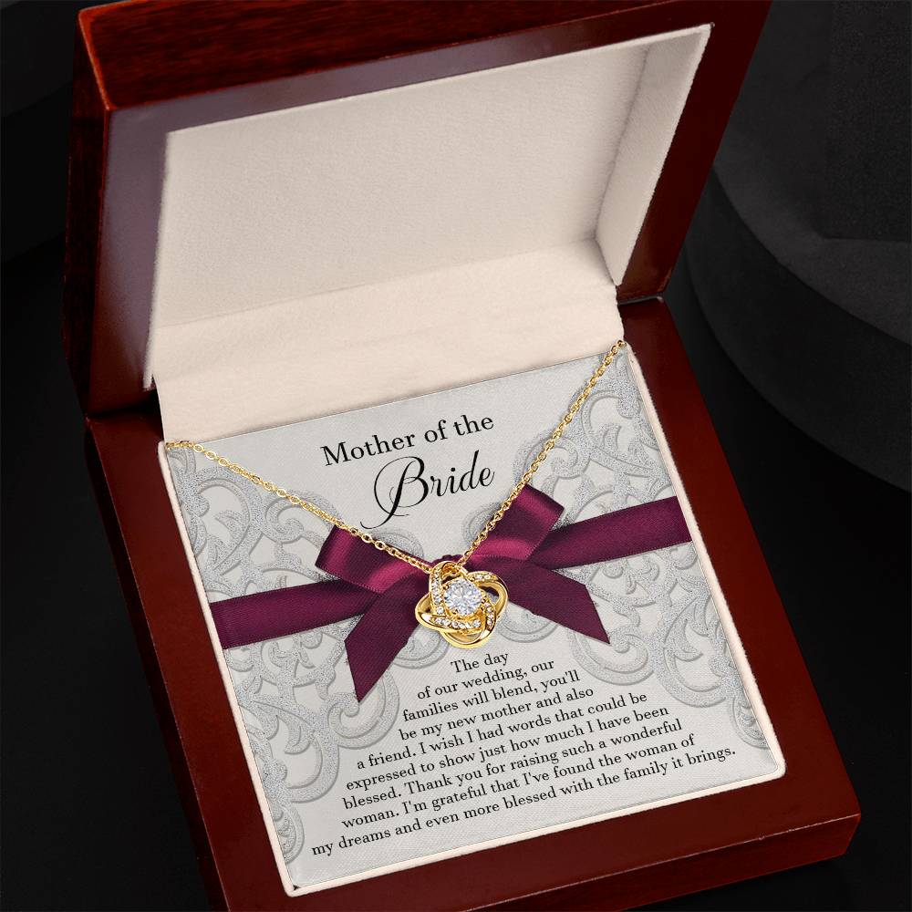 The "To Bride's Mom, My New Mother - Love Knot Necklace" with a gold heart pendant is displayed in an open jewelry box. The box has a message reading "Mother of the Bride" with a sentimental note beneath it. This personalized gift rests on a maroon ribbon, symbolizing the unbreakable bond between mother and daughter.