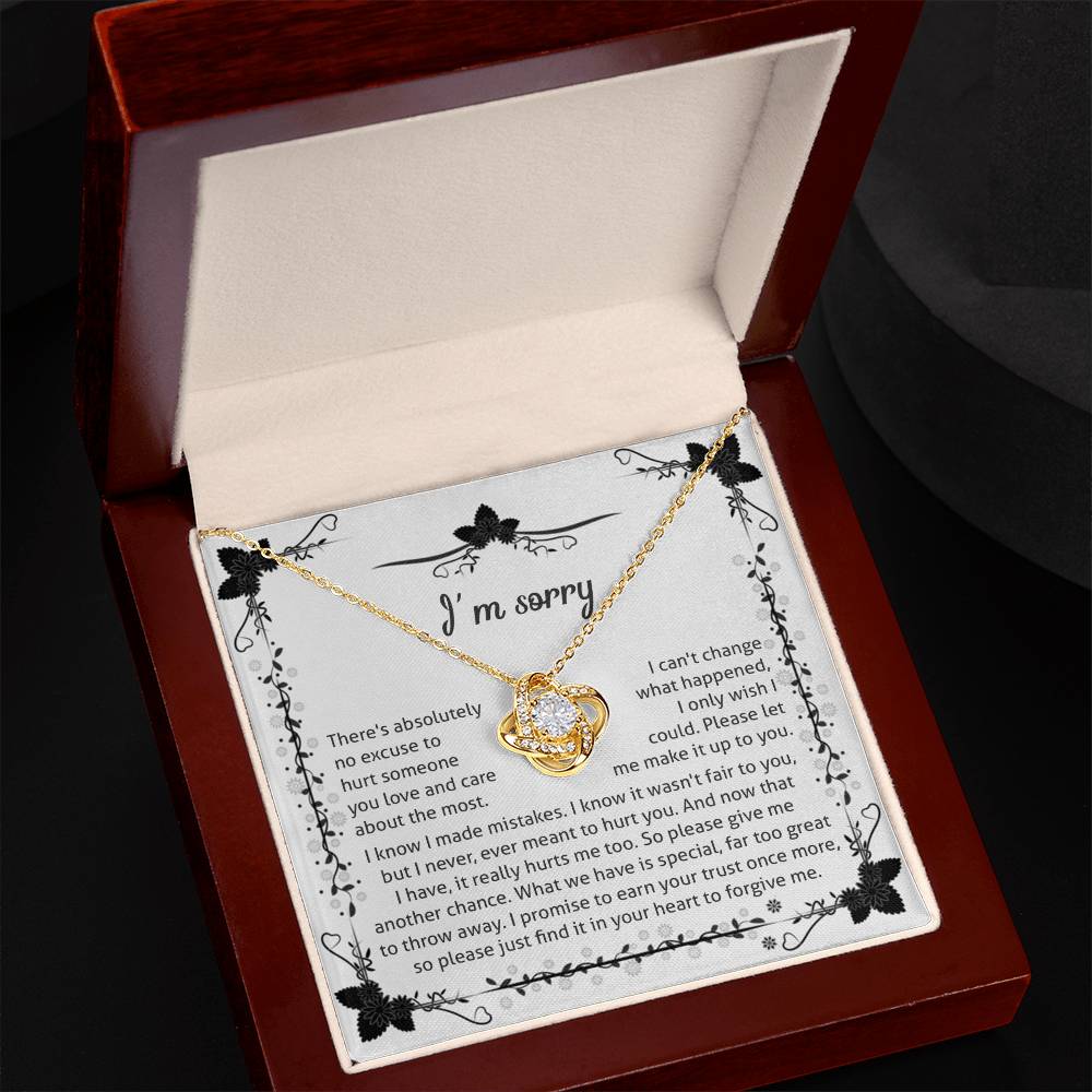 The Sorry-No Excuse - Love Knot Necklace, a gold heart-shaped piece adorned with cubic zirconia crystals, is elegantly displayed inside an open red velvet-lined jewelry box. The lid features an attached note that begins with "I'm sorry" and conveys heartfelt apologies, decorated with floral patterns in the corners—making it an exquisite gift for a loved one.