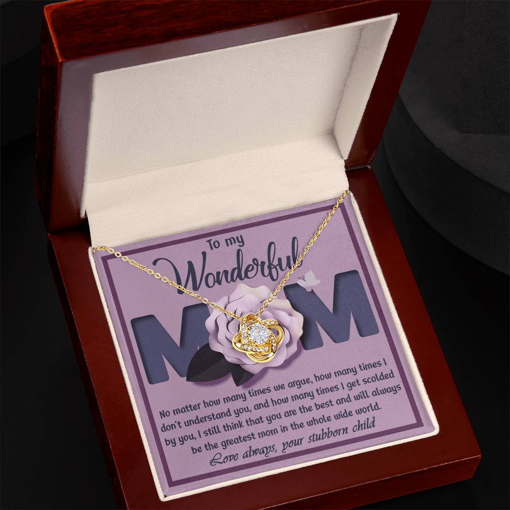 A jewelry box showcases the "To Mom, The Greatest Mom - Love Knot Necklace," featuring a pendant in the shape of a heart adorned with cubic zirconia crystals. It is placed on a card bearing a heartfelt message from a "stubborn child" to "Mom." This personalized gift beautifully expresses unspoken love.