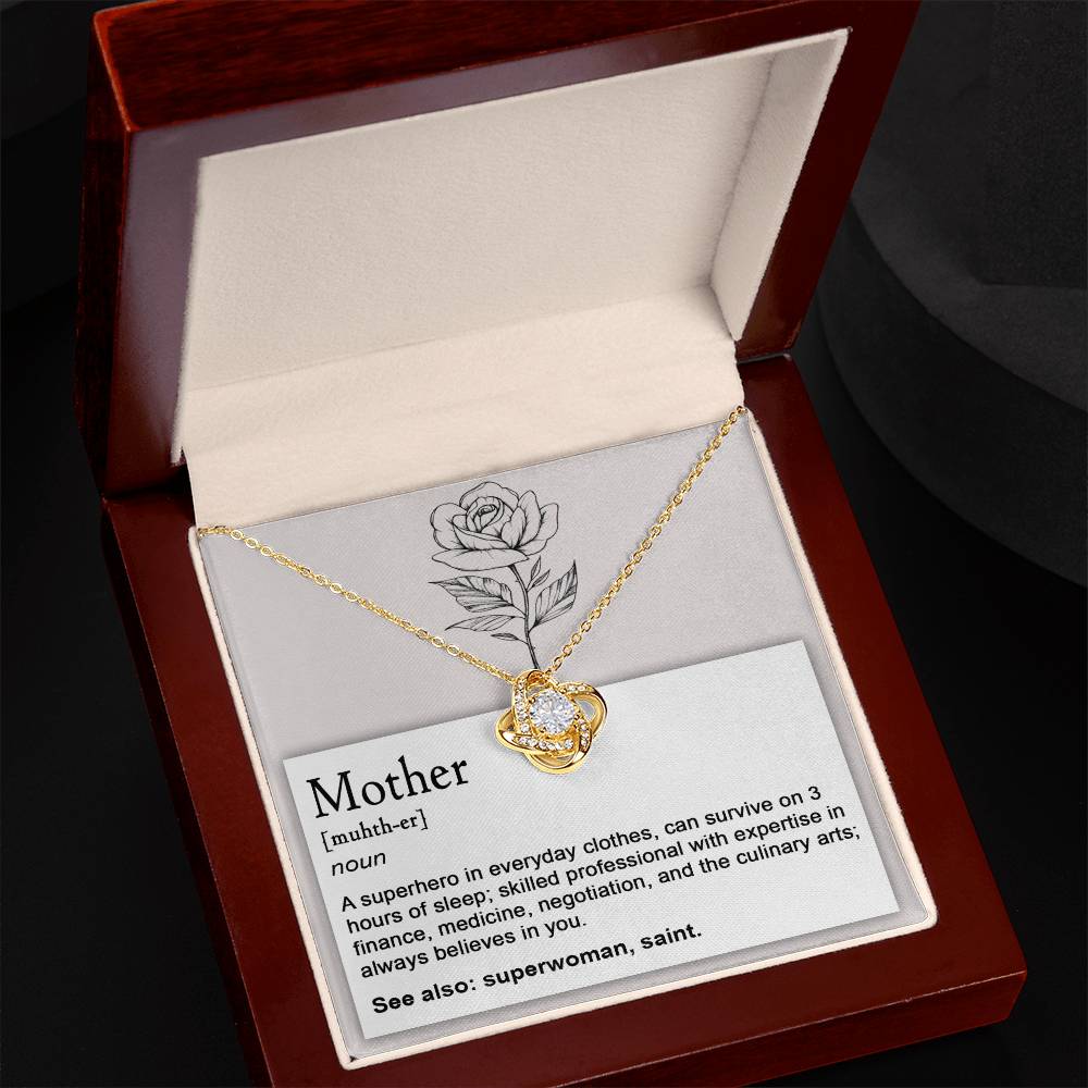 The "To Mom, Superhero - Love Knot Necklace" is adorned with cubic zirconia crystals and comes in a wooden box. It includes a card defining "Mother" as a superhero skilled in multitasking, sleep deprivation, and various skills. The card mentions "superwoman" and "saint," showcasing the necklace's white gold finish.