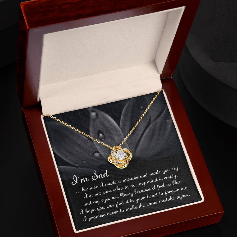 The "Sorry-Feel So Blue - Love Knot Necklace," featuring a stunning knot pendant adorned with cubic zirconia crystals, is displayed over a card with an apologetic message starting with "I'm Sad" and set against a floral background. This personalized gift beautifully combines elegance and heartfelt sentiment.