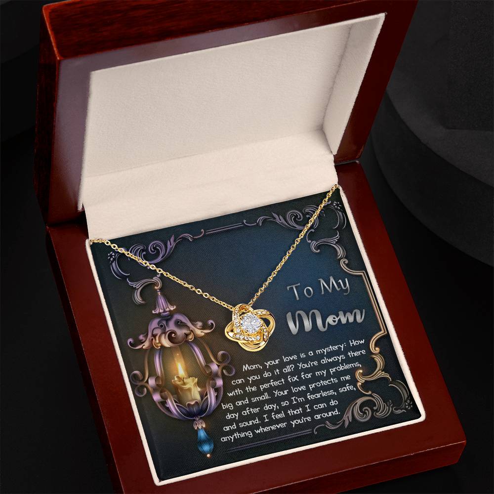 The "To Mom, Love Is Mystery - Love Knot Necklace," featuring a heart-shaped pendant with a gold finish and an inscribed message to a mother, is elegantly displayed in an open jewelry box and adorned with delicate cubic zirconia.