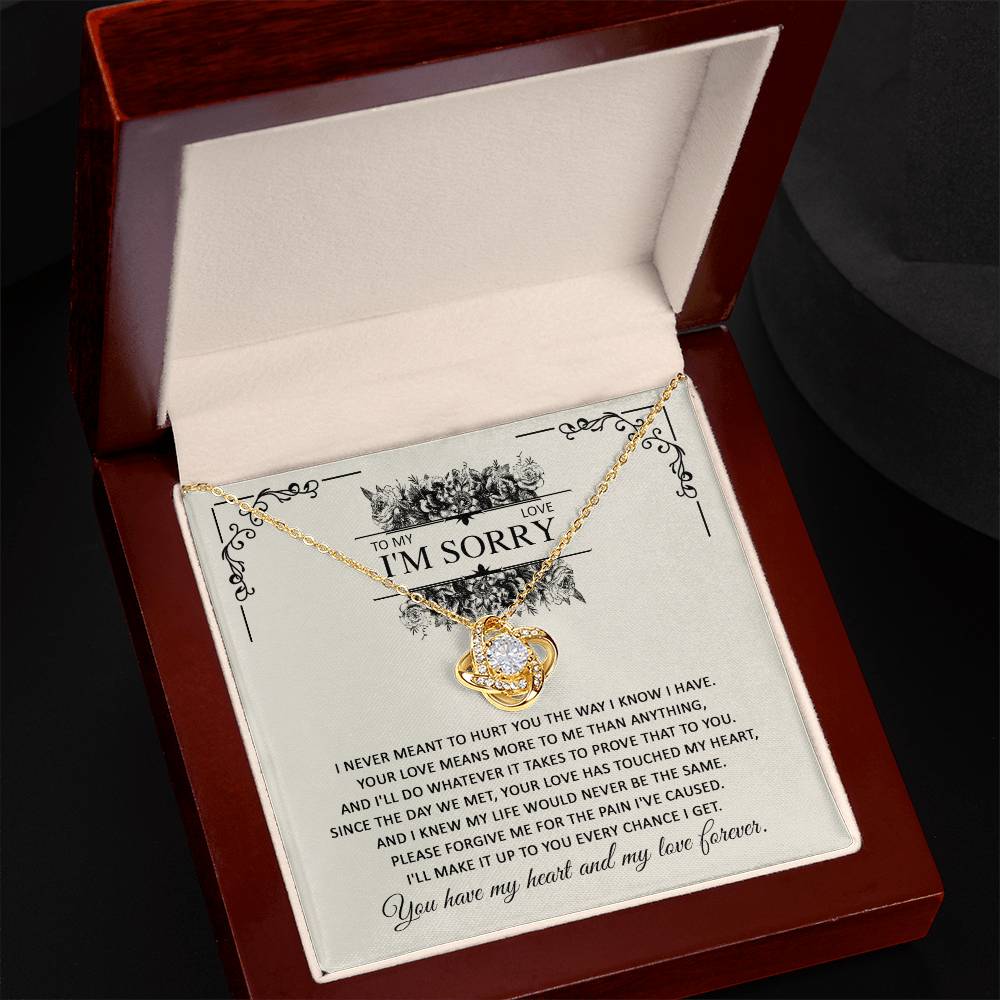 The Sorry-Every Chance Love Knot Necklace, featuring an intertwined heart pendant and adorned with cubic zirconia crystals, is presented in an open, dark red wooden box. The white, padded interior of the box features a heartfelt apology message on the inside lid with the words "I'm Sorry" prominently displayed at the top.