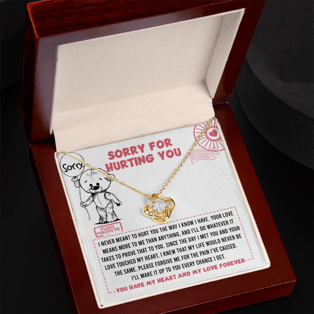 The "Sorry-Every Chance - Love Knot Necklace," featuring a gold finish and a heart pendant adorned with cubic zirconia crystals, is displayed in an open wooden jewelry box. The box lid showcases an illustration of a bear and an apology message.