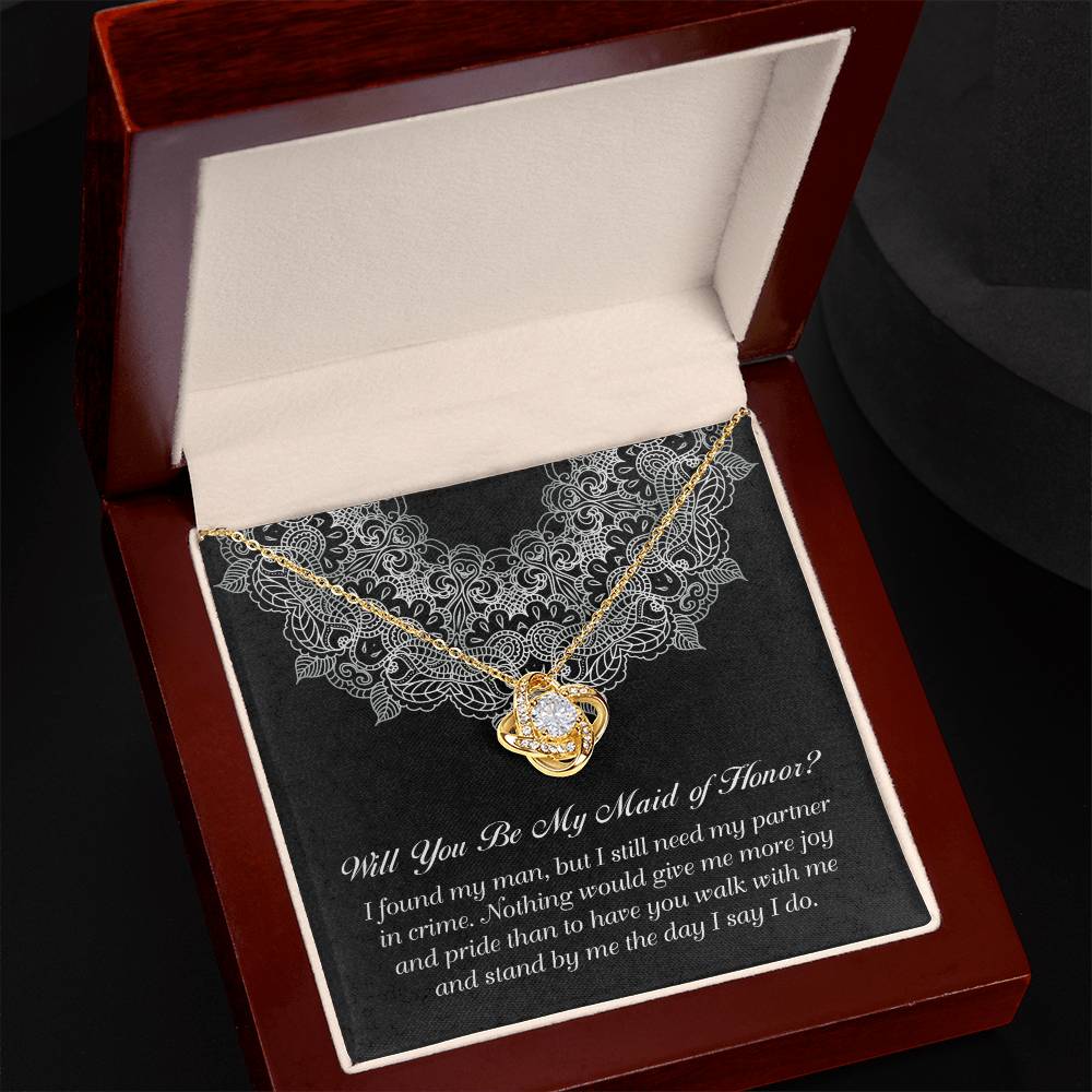 A "To Maid Of Honor, Partner In Crime - Love Knot Necklace" featuring an intertwined heart pendant, elegantly presented in a wooden box with a white satin lining and a decorative black background. Below the necklace is the heartfelt message, "Will you be my maid of honor?" along with an additional sentimental message about friendship and partnership.