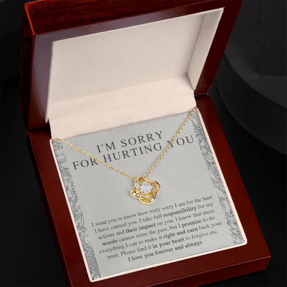 The Sorry-Do Everything - Love Knot Necklace, adorned with cubic zirconia crystals, is showcased in an open velvet-lined jewelry box. Behind the necklace, an apology message is visible. The box features a rich, dark wooden exterior and a cream-colored fabric interior.