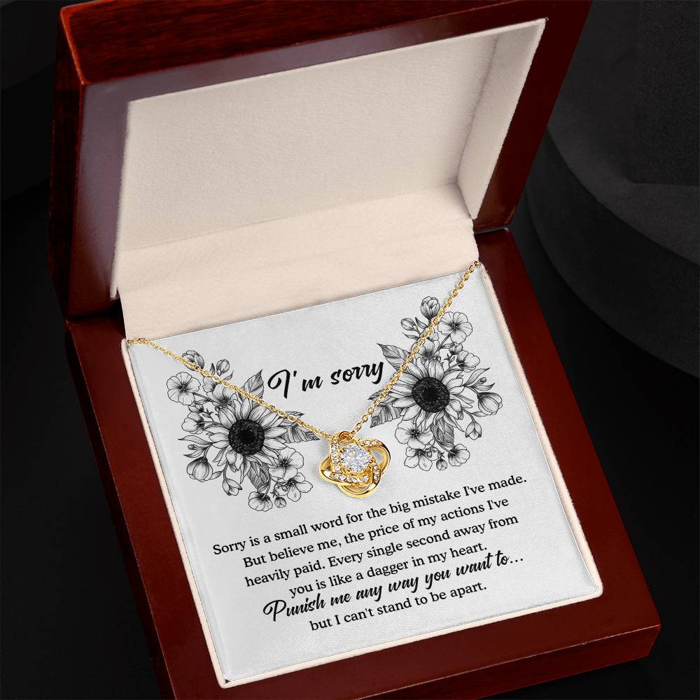 A jewelry gift box containing a Sorry-Small Word Love Knot Necklace, featuring two intertwined hearts with a gold finish, each adorned with a small diamond. Inside the lid, an apology message is printed amidst black and white floral illustrations, expressing regret and the pain of being apart.