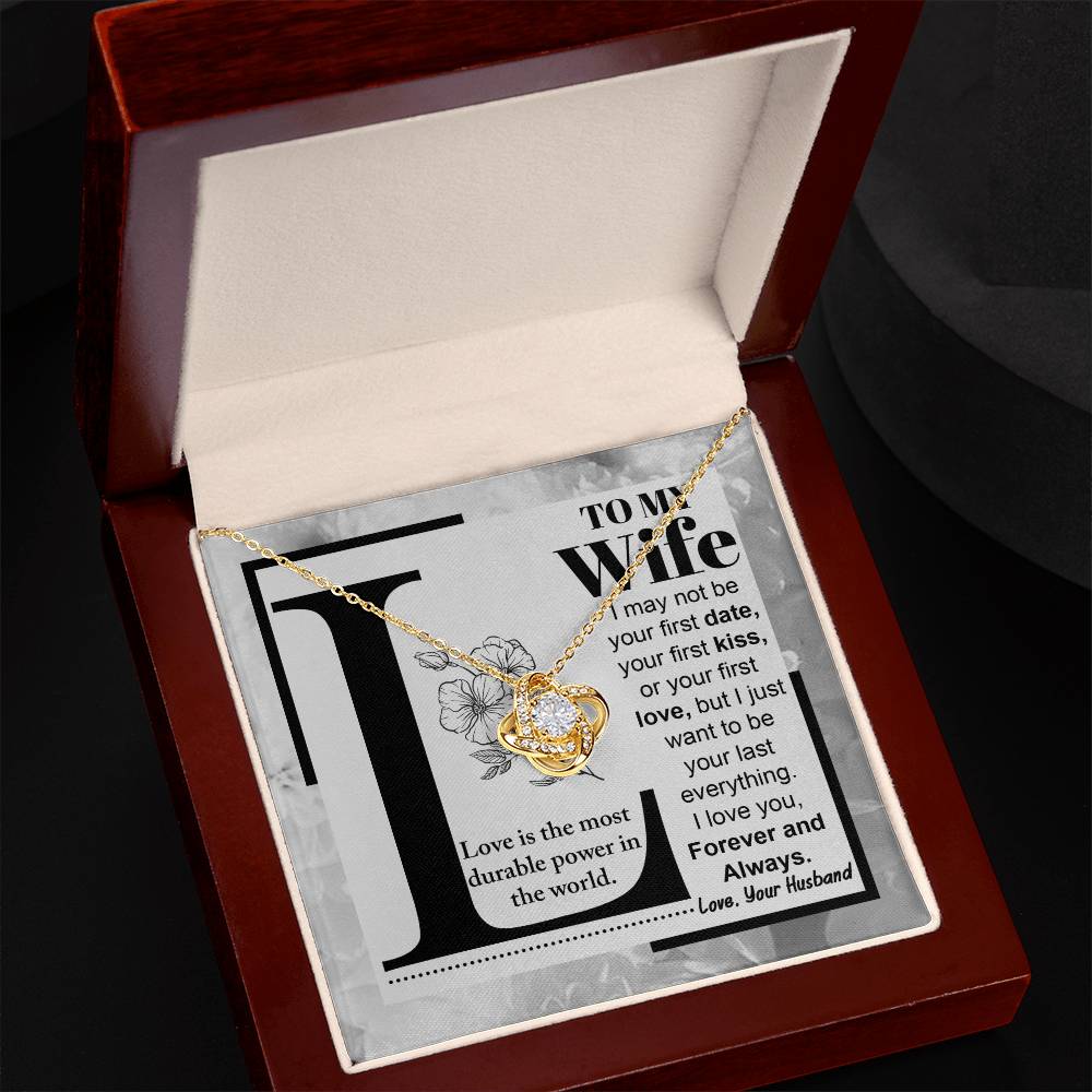 A "to Wife, Love Is - Love Knot Necklace" with a knot design is displayed on a card featuring a printed message to a wife about lasting love, signed from a husband. The piece sparkles with cubic zirconia crystals and boasts an elegant gold finish.