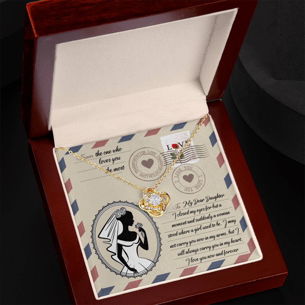 The "To Daughter Wedding, In My Arms - Love Knot Necklace," adorned with cubic zirconia crystals, is exquisitely displayed in an open wooden box. Inside, a printed message showcases an illustration of a father and daughter dancing on a postcard-themed background, making it the perfect personalized gift.