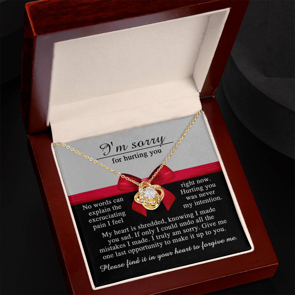 The Sorry-Made You Sad - Love Knot Necklace, featuring a heart pendant, is displayed in an open wooden box. Inside the box, a printed message reads, "I'm sorry for hurting you," accompanied by an apology note. The necklace shines with its white gold finish and is adorned with cubic zirconia crystals that catch the light beautifully.